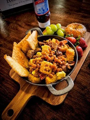 A full English breakfast in a cast-iron skillet — beans, sausage, bacon and fried bread on a wooden board, with a bottle of brown sauce behind
